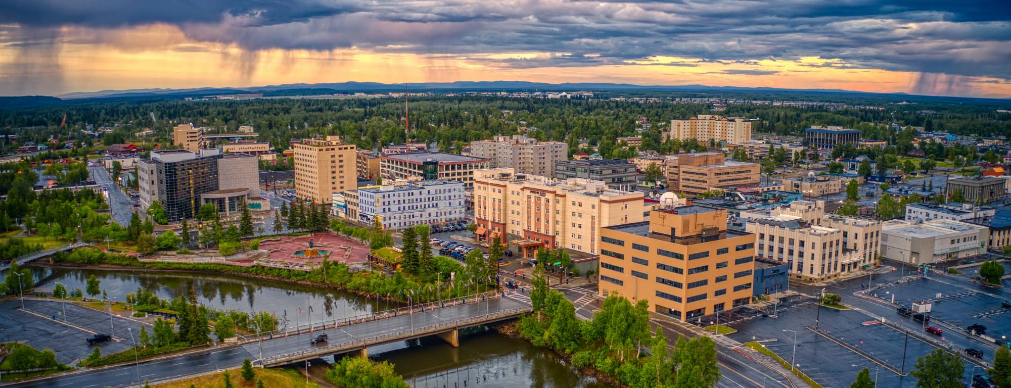 Downtown Fairbanks and the Chena River under storm clouds and a sunset glow, with hills on the horizon