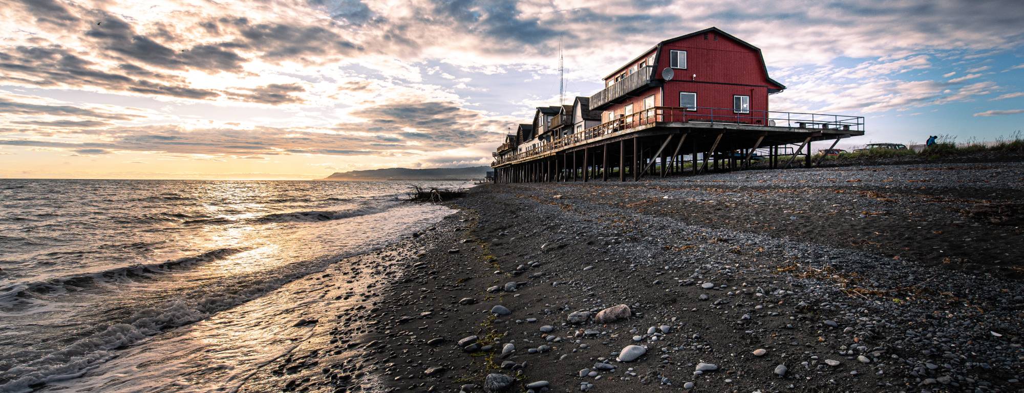 A red building on pilings above a dark pebble beach in Homer, Alaska, with waves and a cloudy sunset sky over the water